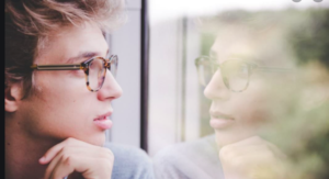 Young man with glasses looking through a window at his reflection